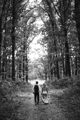 Fototapeta premium Black and white foto the newlyweds walk hand in hand along the path in the forest. Back view. Bride and groom enjoying romantic moment together on wedding day. The concept of youth, love and lifestyle