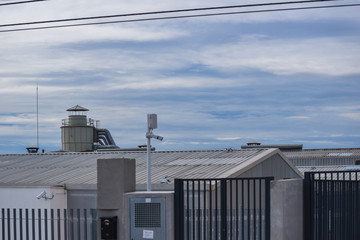 view of an industrial environment with a ship with a large gray roof in the foreground with a fence, security cameras and a liquid tank at the end with all gray pipes on a cloudy day