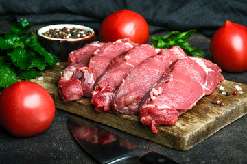 Fresh raw beef, cut into steaks on a wooden board with vegetables, herbs and spices on a wooden board on a dark background.