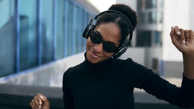 Pretty afro girl happy smiling. Stunning african woman with light-brown skin relaxing in headphones and funny dancing on modern city background. Wearing black sweater and stylish black sunglasses