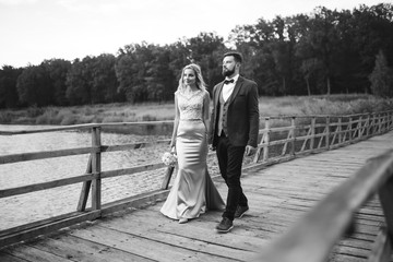 Black and white foto stylish couple of newlyweds posing on a bridge on wedding day. Handsome bearded groom admires and kisses pretty bride. Together. The concept of youth, love, fashion and lifestyle.