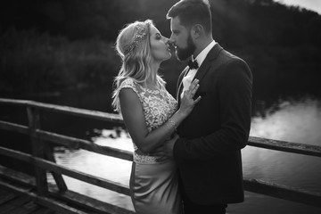 Black and white foto stylish couple of newlyweds posing on a bridge on wedding day. Handsome bearded groom admires and kisses pretty bride. Together. The concept of youth, love, fashion and lifestyle.