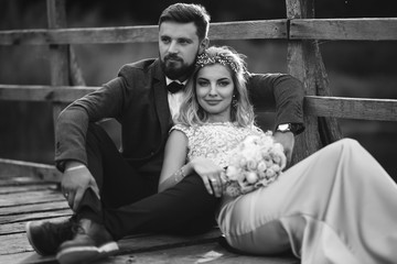 Black and white foto stylish couple of newlyweds posing on a bridge on wedding day. Handsome bearded groom admires and kisses pretty bride. Together. The concept of youth, love, fashion and lifestyle.