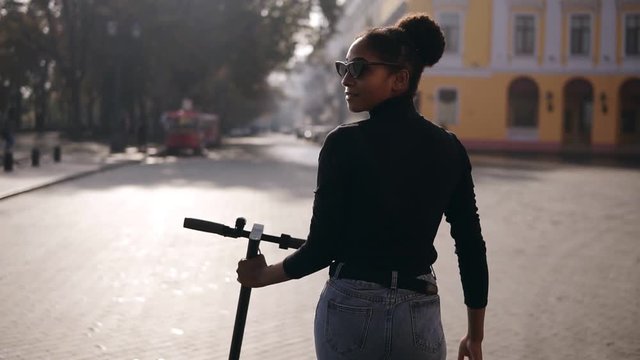 Rare view footage of mix raced woman walking along the city street. Girl in black coat and sunglasses walking with electric kick scooter by the morning city, looking back over the shoulder. Slow