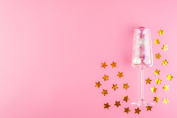 A glass of champagne is filled with pink and gold shiny decorations in the form of balls on a pink pastel background