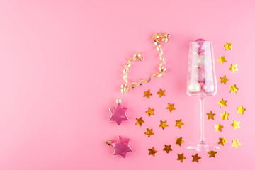 A glass of champagne is filled with pink and gold shiny decorations in the form of balls on a pink pastel background