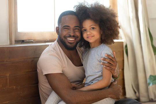 Smiling biracial dad and preschooler daughter hug looking at camera
