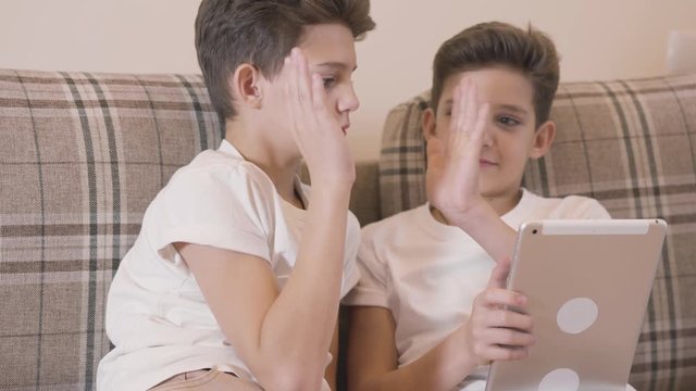 Close-up portrait of two Caucasian twins playing video games at tablet and giving high five. Brothers spending free time together indoors. Siblings resting at home.
