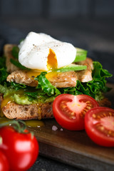 Fototapeta na wymiar Delicious sandwich with avocado and poached egg, cherry tomatos and arugula on a dark background.