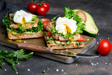 Delicious sandwich with avocado and poached egg, cherry tomatos and arugula on a dark background.