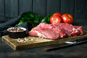 Fresh raw beef, cut into steaks  with vegetables, herbs and spices on a wooden board on a dark background.