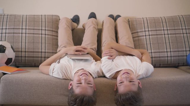 Two Caucasian twin brothers laying laying upside down on sofa, looking at camera and laughing. Cheerful siblings resting together at home. Modern kids having fun indoors.