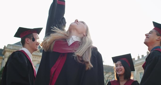 Portrait shot of the young beautiful blond Caucasian girl, master graduate smiling joyfully to the camera and tossing up in the air her cap. Outdoors.