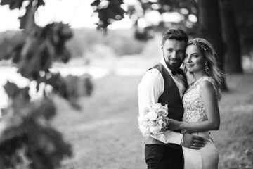 Sensual Black and white foto of bride and groom. Stylish couple of happy newlyweds posing in the park on their wedding day. Handsome bearded groom admires and kisses pretty bride. Together.