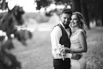Sensual Black and white foto of bride and groom. Stylish couple of happy newlyweds posing in the park on their wedding day. Handsome bearded groom admires and kisses pretty bride. Together.