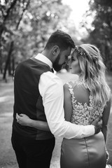 Sensual Black and white foto of bride and groom. Stylish couple of happy newlyweds posing in the park on their wedding day. Handsome bearded groom admires and kisses pretty bride. Together.