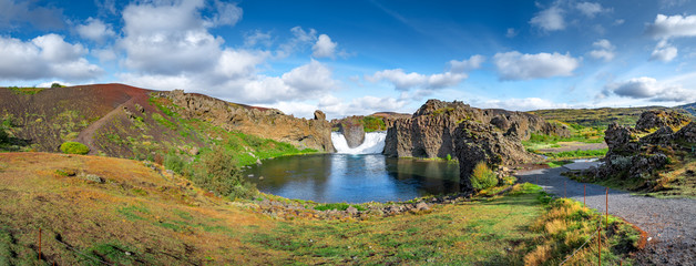 Epic aerial drone panorama view flying over landscape of Hjalparfoss waterfall and lagoon on a sunny day.