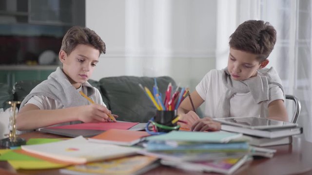 Two smart Caucasian twin brothers drawing at home with colorful pencils. Schoolboys doing homework together. Creative siblings sitting at the table and painting.