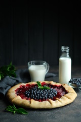 Homemade backing. Delicious galette with plums and blueberries with cup of milk on a dark background copy space.