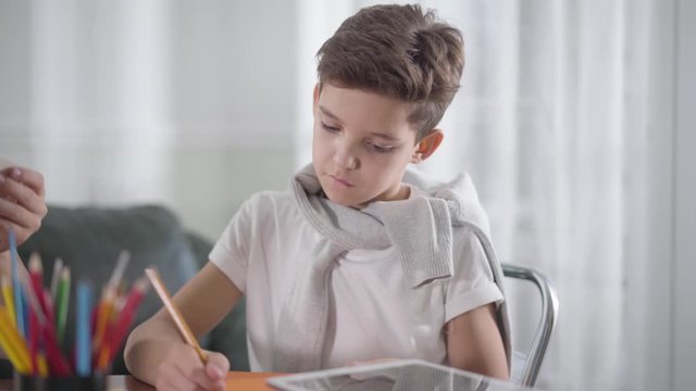 Portrait of concentrated Caucasian boy sitting at the table and drawing or writing in notebook. Schoolboy doing homework at home. Education, intelligence, bright mind, generation z
