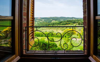 Fototapeta na wymiar Open window with a view of the garden in Tuscany rural landscape. Countryside, small medieval town, cypresses trees, green field, sun light and cloud. Italy, Europe.