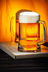 Two Light Beer Mugs with White Foam on Dark Wooden Table and Yellow Background