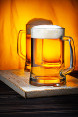 Two Light Beer Mugs with White Foam on Dark Wooden Table and Yellow Background