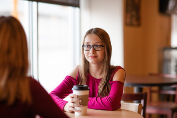 Happy beautiful teenager girls, two sisters in glasses, sitting in cafe drinking coffee and have conversation. smiling and taking 
