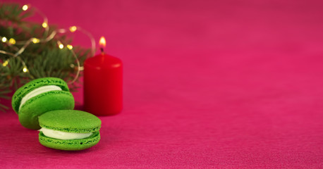 Green macaron with fondant on red paper background. Nearby is a Christmas tree branch with a garland and a burning red candle. Close-up, copy space for text. Postcard or banner concept.