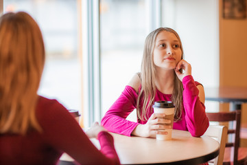 Happy beautiful teenager girls, two sisters, sitting in cafe drinking coffee and have conversation. smiling and taking 