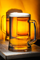 Two Light Beer Mugs with White Foam on Dark Wooden Table and Yellow Background