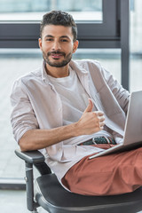 smiling businessman in casual clothes sitting on office chair, showing thumb up and looking at camera
