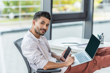 young, confident businessman holding laptop and smartphone with blank screen while looking at camera