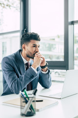 thoughtful businessman looking away while sitting at workplace and holding folded hands near face