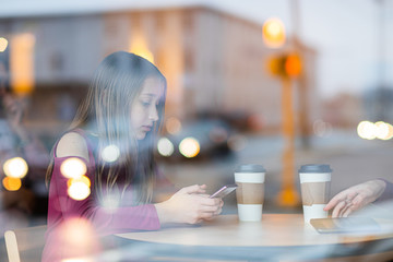 Cute, sad, serious, beautiful teenager girl sitting in cafe drinking coffee and chatting on mobile phone. picture through window