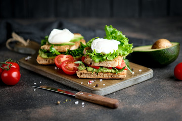 Delicious sandwich with avocado and poached egg, with green leaves and tomatoes on a dark background. Healthy breakfast.