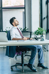 thoughtful call center operator looking away while sitting at workplace near laptop and headset