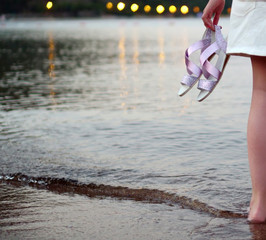 A girl stands at sea, holds shoes in her hand, against the backdrop of lights from the shore
