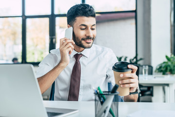 smiling businessman holding coffee to go while talking on smartphone