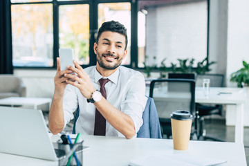 cheerful businessman holding smartphone while sitting at workplace near laptop and disposable cup