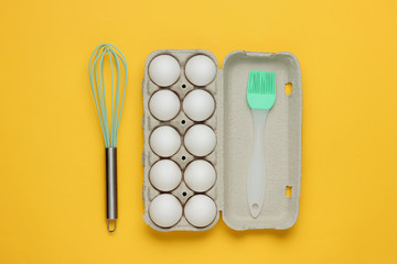 The concept of cooking. Cardboard tray of eggs, kitchen tools (whisk, brush) on yellow background. Top view