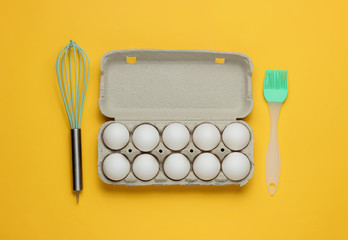 The concept of cooking. Cardboard tray of eggs, kitchen tools (whisk, brush) on yellow background. Top view
