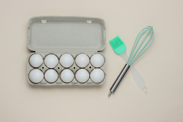 The concept of cooking. Cardboard tray of eggs, kitchen tools (whisk, brush) on white  background. Top view