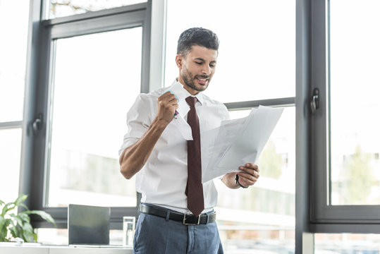 dissatisfied businessman crumpling paper while standing near window in office - Powered by Adobe