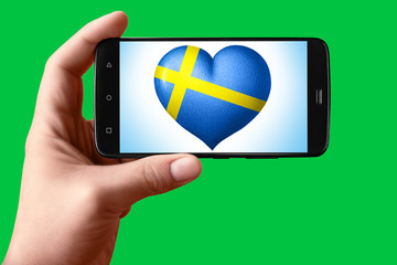Sweden flag in the shape of a heart on the phone screen. A smartphone in hand shows a flag heart on a chroma key background.
