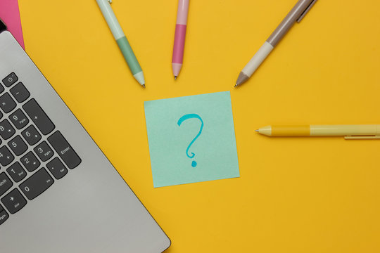 Laptop with pens and memo piece of paper with question mark on yellow background. Top view