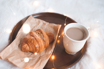Tasty croissant with cup of coffee over glowing lights in room closeup. Good morning.