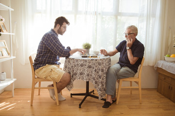 Young man and elder old man playing chess board game together at home.