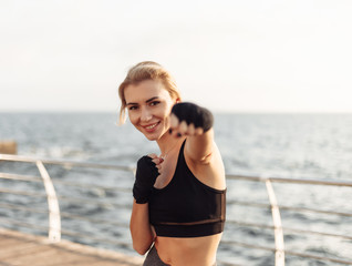 Young woman kickboxer in sportwear with wrapped hands in bandages trains on the beach at sunrise. Morning training fighter