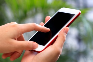 Hand holding smartphone and forefinger touching on blank screen in green garden.